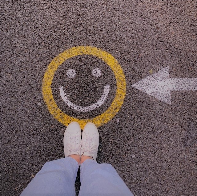 Das Bild zeigt einen auf der Straße aufgemalten Smiley, vor dem eine Person steht, von der nur die Füße zu sehen sind (Quelle: jacqueline-munguia-1pAwJiCD60c/ Unsplash) Das Bild zeigt einen auf der Straße aufgemalten Smiley, vor dem eine Person steht, von der nur die Füße zu sehen sind