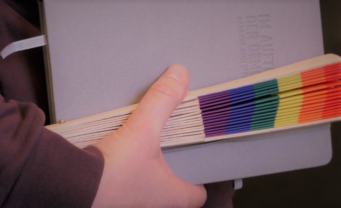 Hand mit Notizbuch und eingeklapptem Fächer in Regenbogenfarben Hand mit Notizbuch und eingeklapptem Fächer in Regenbogenfarben