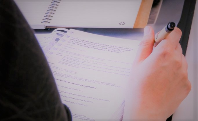 Blick von hinten auf eine Frau an einem Schreibtisch. Sie hat einen Stift in der Hand und vor ihr liegen ein Notizbuch und einen Gesetzestext. Blick von hinten auf eine Frau an einem Schreibtisch. Sie hat einen Stift in der Hand und vor ihr liegen ein Notizbuch und einen Gesetzestext.