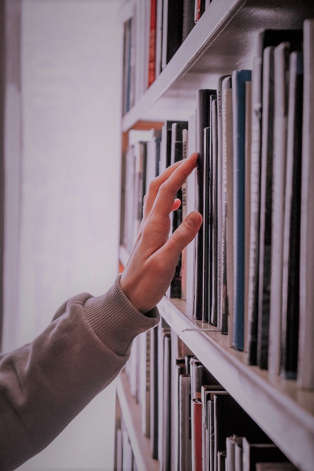 Das Bild zeigt einen Hand, die über Buchtitel in einem Bücherregal streicht (Quelle: guzel-maksutova-B30XL_m3fso/unsplash) Das Bild zeigt einen Hand, die über Buchtitel in einem Bücherregal streicht