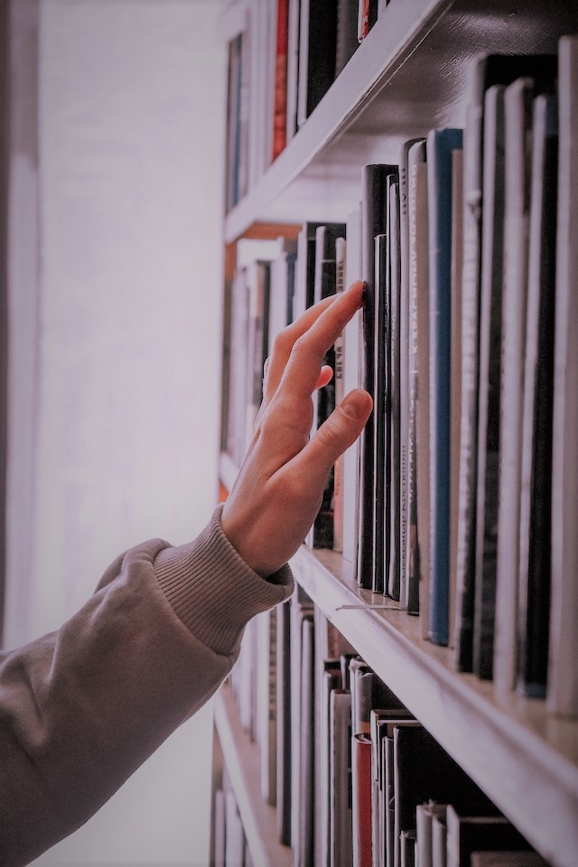 Das Bild zeigt einen Hand, die über Buchtitel in einem Bücherregal streicht