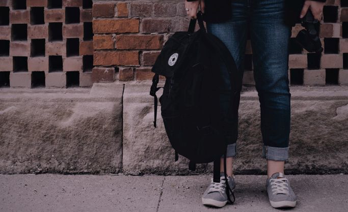 Das Bild zeigt eine Person mit einer Tasche vor einer Mauerwand. (Quelle: Scott Webb - unsplash) Das Bild zeigt eine Person mit einer Tasche vor einer Mauerwand.