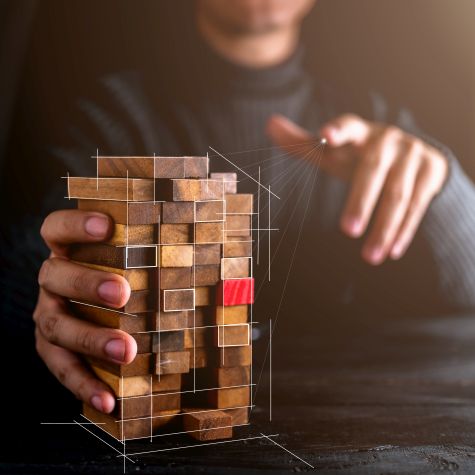 Die Abbildung zeigt einen Geschäftsmann, der einen Holzblock untersucht, bei dem ein einzelner Baustein - als potenzielle Schwachstelle - eine rote Farbe hat (Quelle: whyframestudio / istock / Getty Images Plus) Die Abbildung zeigt einen Geschäftsmann, der einen Holzblock untersucht, bei dem ein einzelner Baustein - als potenzielle Schwachstelle - eine rote Farbe hat