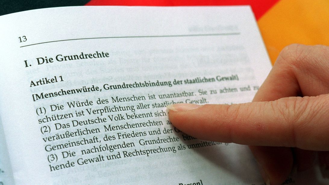 The photograph, taken on April 19, 1999, shows a female index finger pointing to Article 1 of an open edition of the Basic Law. A German flag can be seen in the background.